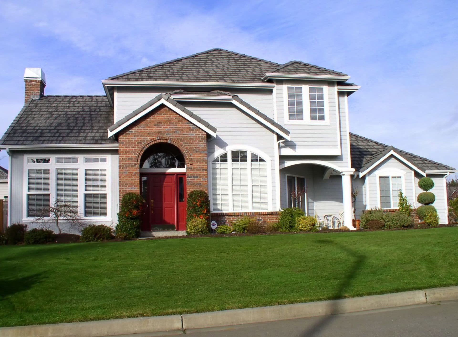 Two-story suburban home with white and brick exterior, red front door with arched entry, attached garage, manicured lawn, and landscaping