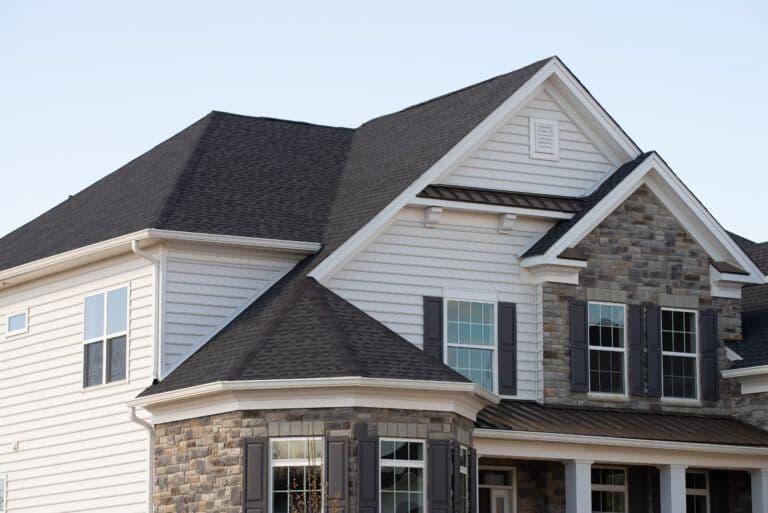 Two-story residential home with gray asphalt shingle roof, white vinyl siding on left section, stone veneer on right facade, and dark shutters framing multiple windows