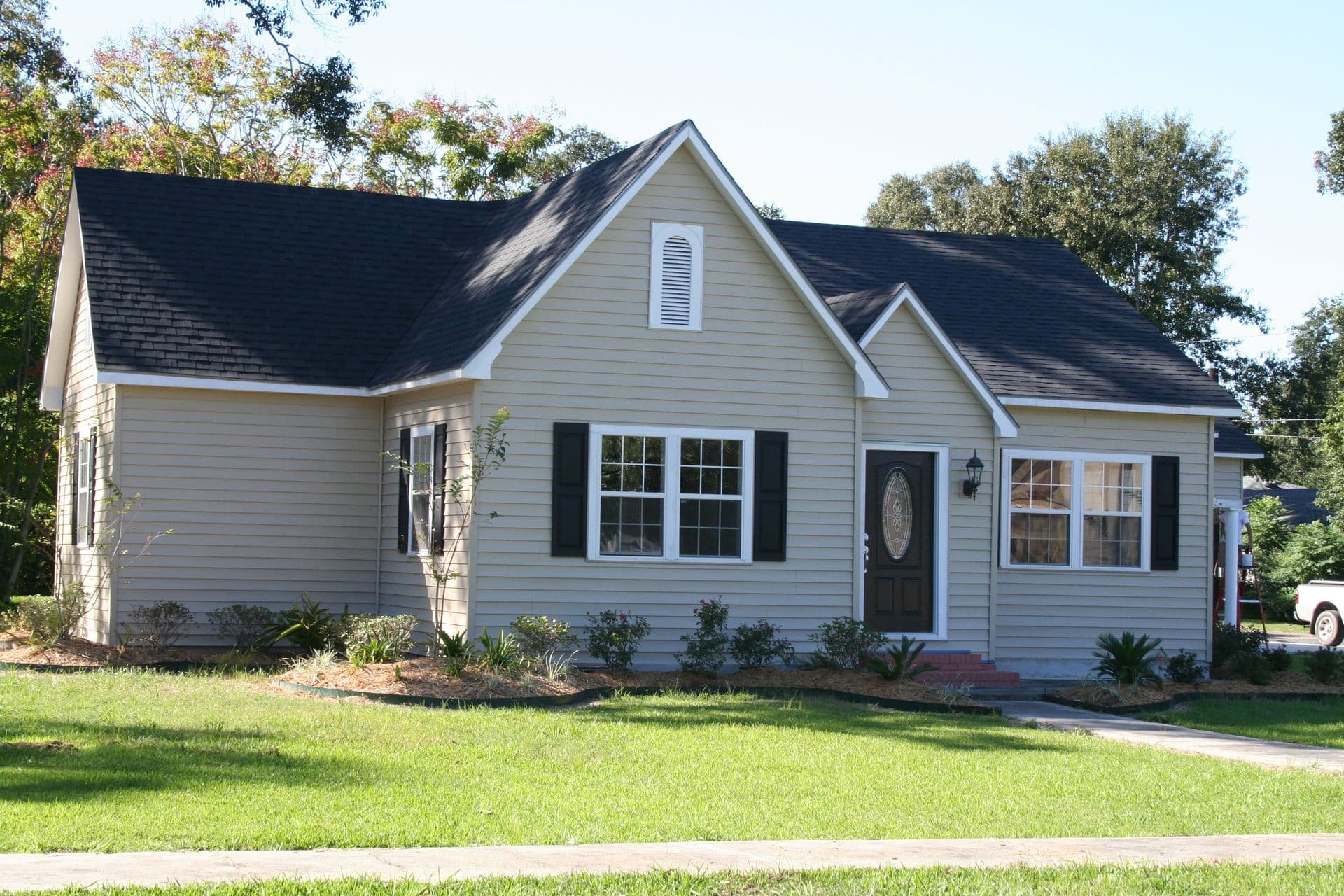 Single-story white house with dark roof, two gable peaks, black shutters, front porch with dark door, and landscaped lawn surrounded by mature trees