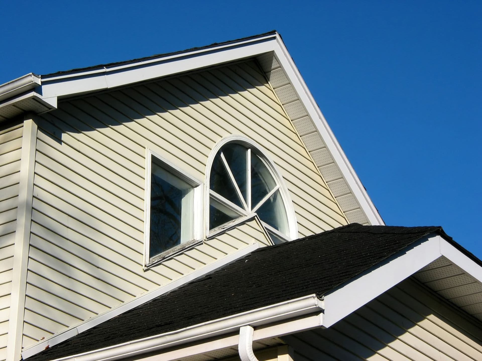 Cream-colored two-story house with oval window in gable, black roof trim, and white fascia against clear blue sky