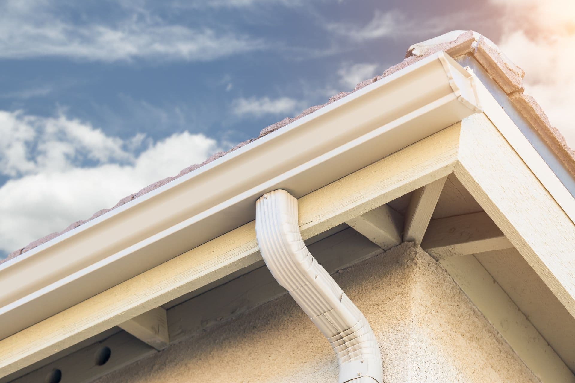 Roof eave with white gutter and downspout against blue sky with clouds