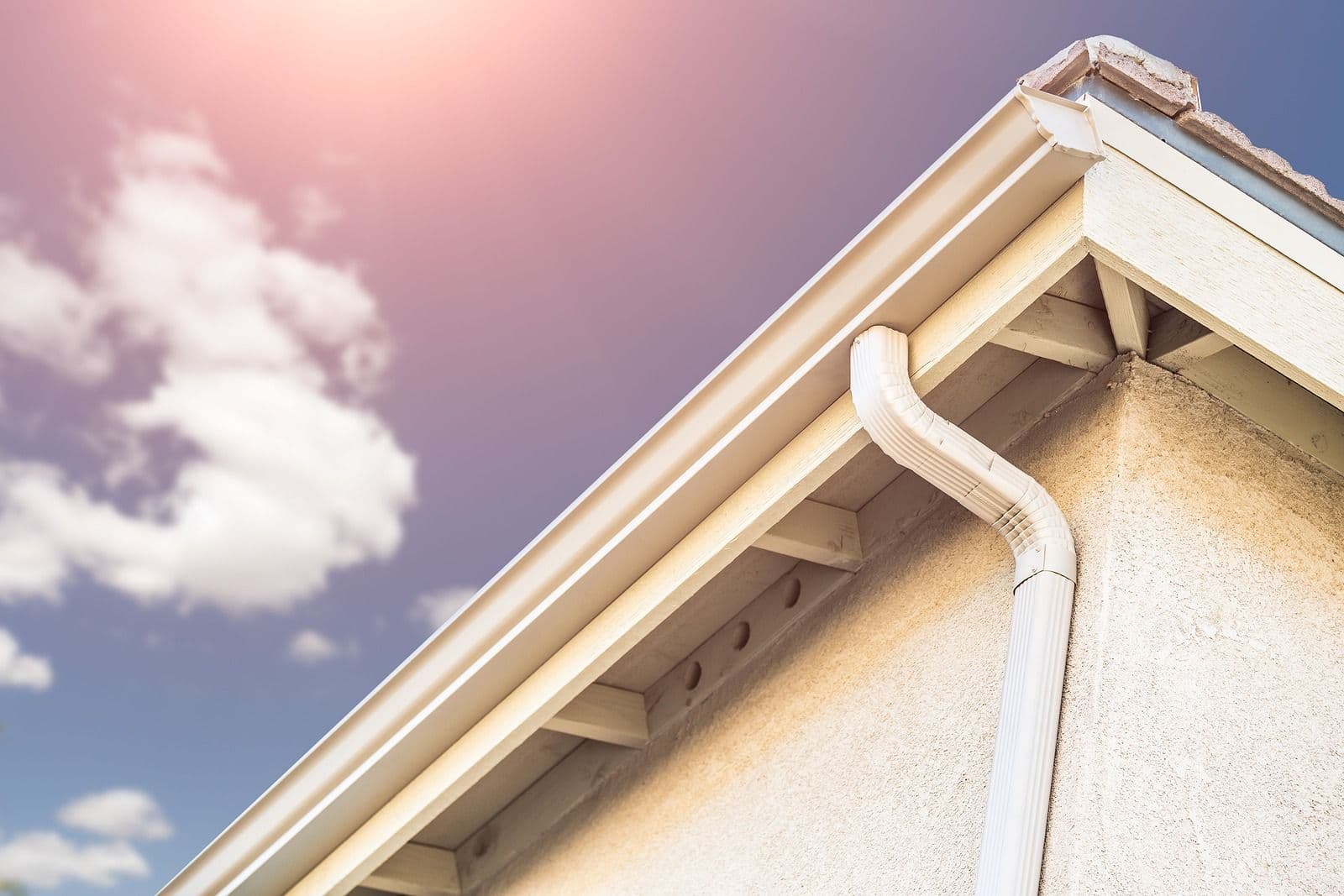 House eave and gutter system with white downspout against blue sky with clouds