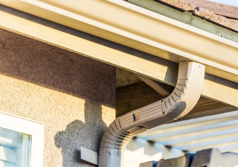 Guttering and downpipe system installed on residential building exterior showing cream-colored fascia and brown corrugated drainage pipe attached to stone wall
