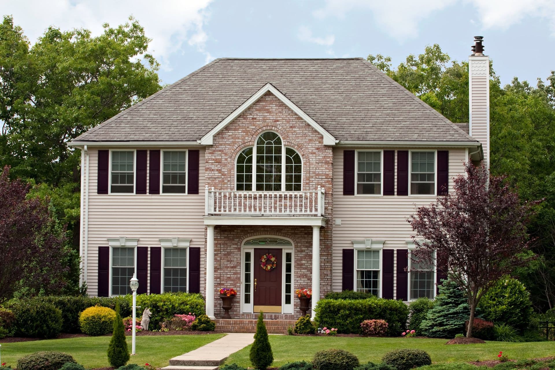 Two-story brick and siding house with arched windows, burgundy shutters, second-floor balcony, front porch with flowering planters, and manicured landscaping