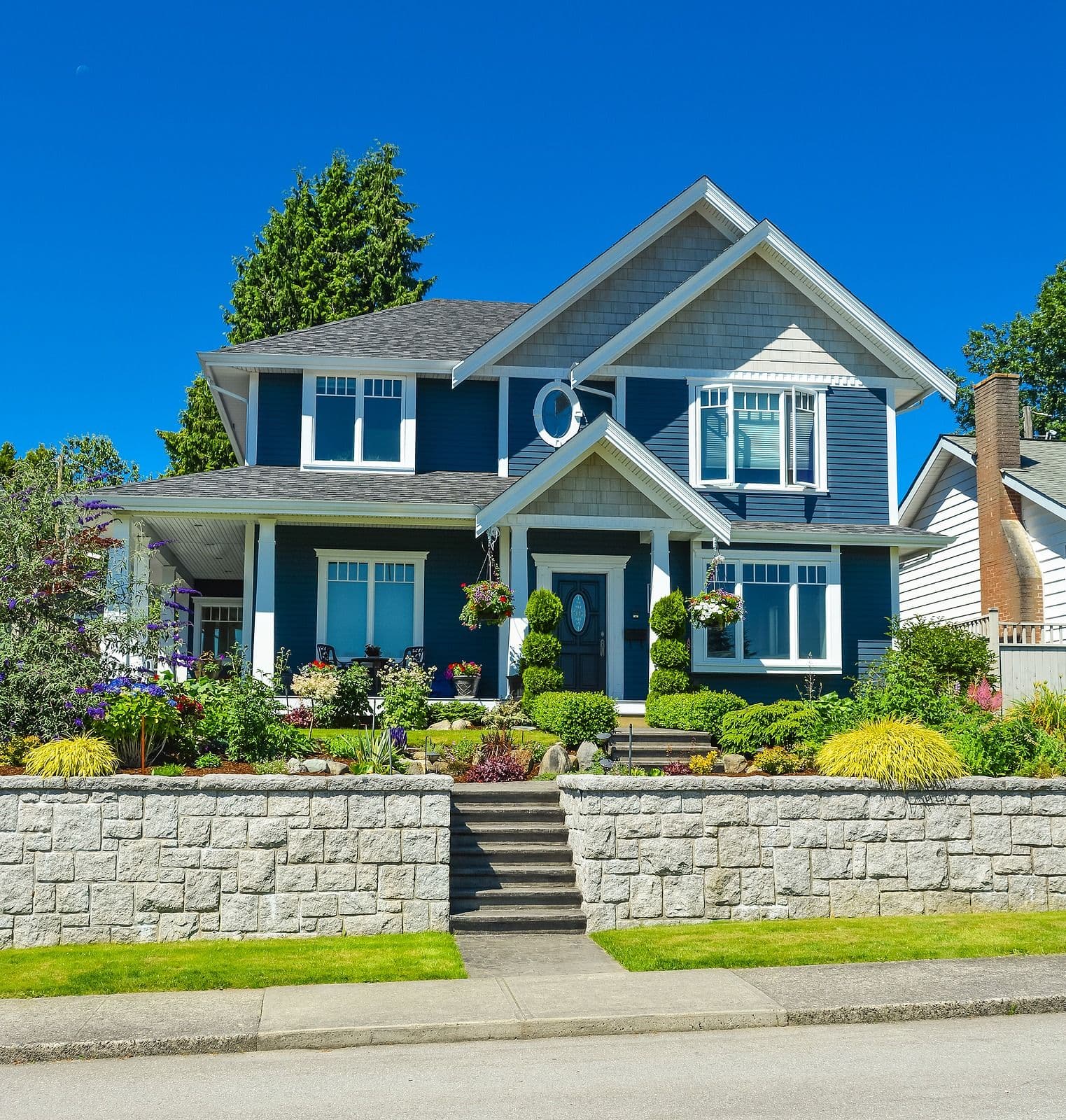Blue two-story house with white trim, stone front wall, and landscaped garden featuring purple flowers and yellow ornamental grasses under a clear blue sky