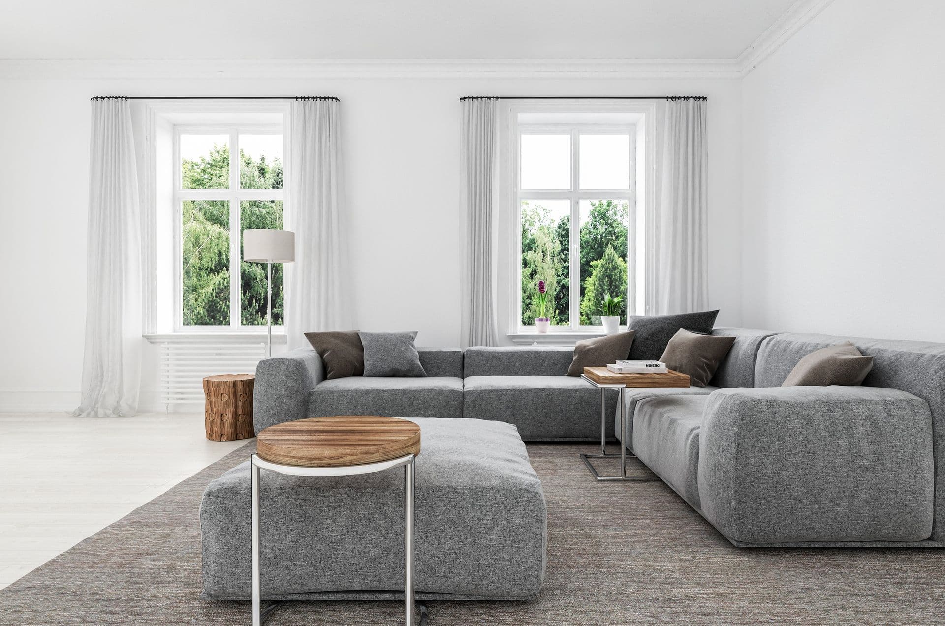 Bright modern living room with gray sectional sofa, wood and metal round coffee table, floor lamp, and two large windows dressed with white curtains overlooking green trees