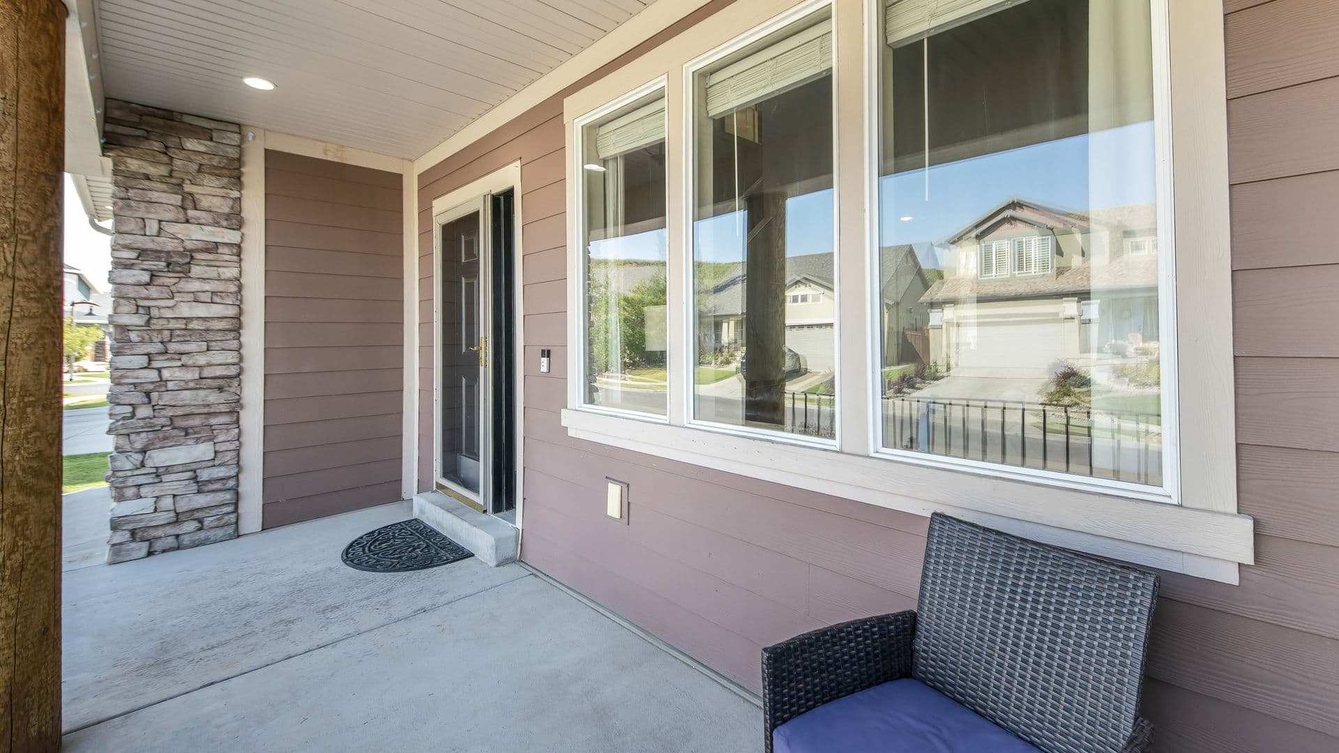 Front porch entry with stone accent wall, dark wooden door, large windows overlooking residential neighborhood, wicker seating, and concrete flooring