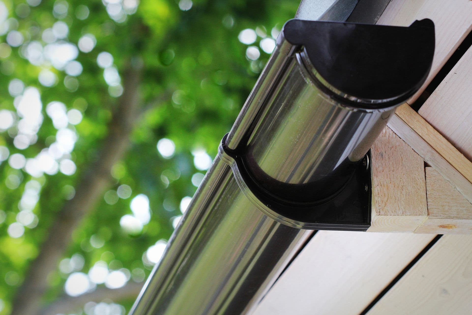 Downspout and gutter system installed on house exterior with green foliage visible in background