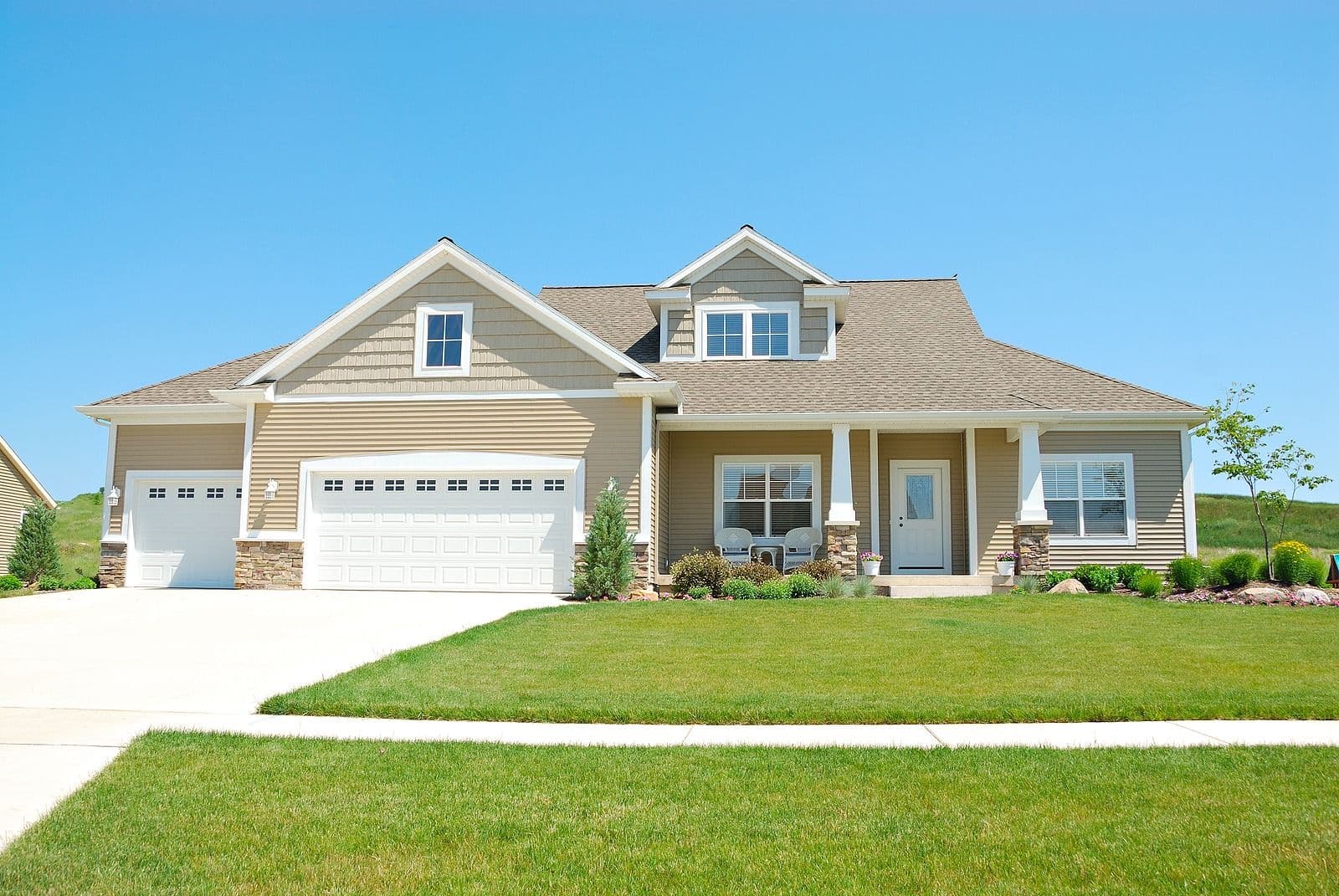 Beige single-story home with two-car garage, front porch with white columns, dormers, and manicured lawn under clear blue sky