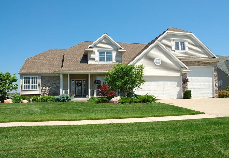 Two-story suburban home with white vinyl siding, brick accents, front porch, and two-car garage, surrounded by manicured lawn and landscaping