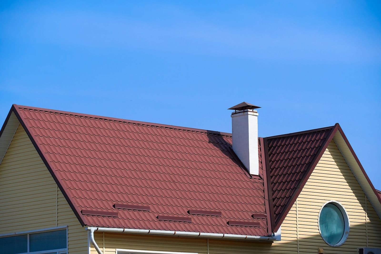 House with burgundy metal tile roof and white chimney against clear blue sky