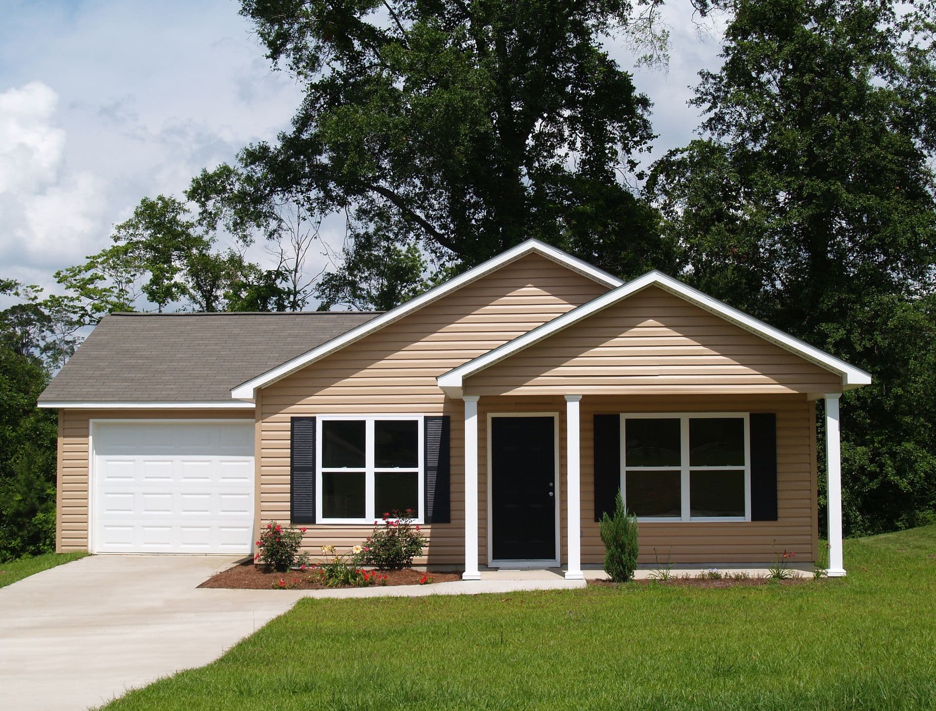 Single-story brown siding house with white-trimmed front porch, garage, and landscaping surrounded by mature trees