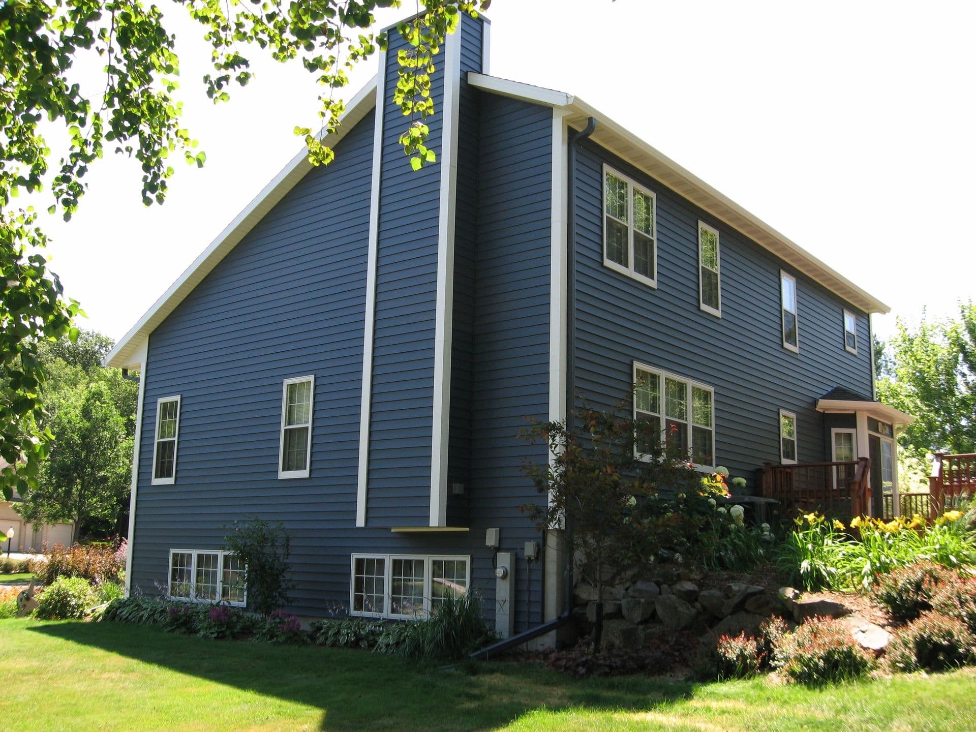 Contemporary blue-gray two-story house with white-trimmed windows, elevated foundation, wooden deck on right side, and landscaped yard with mature trees and shrubs