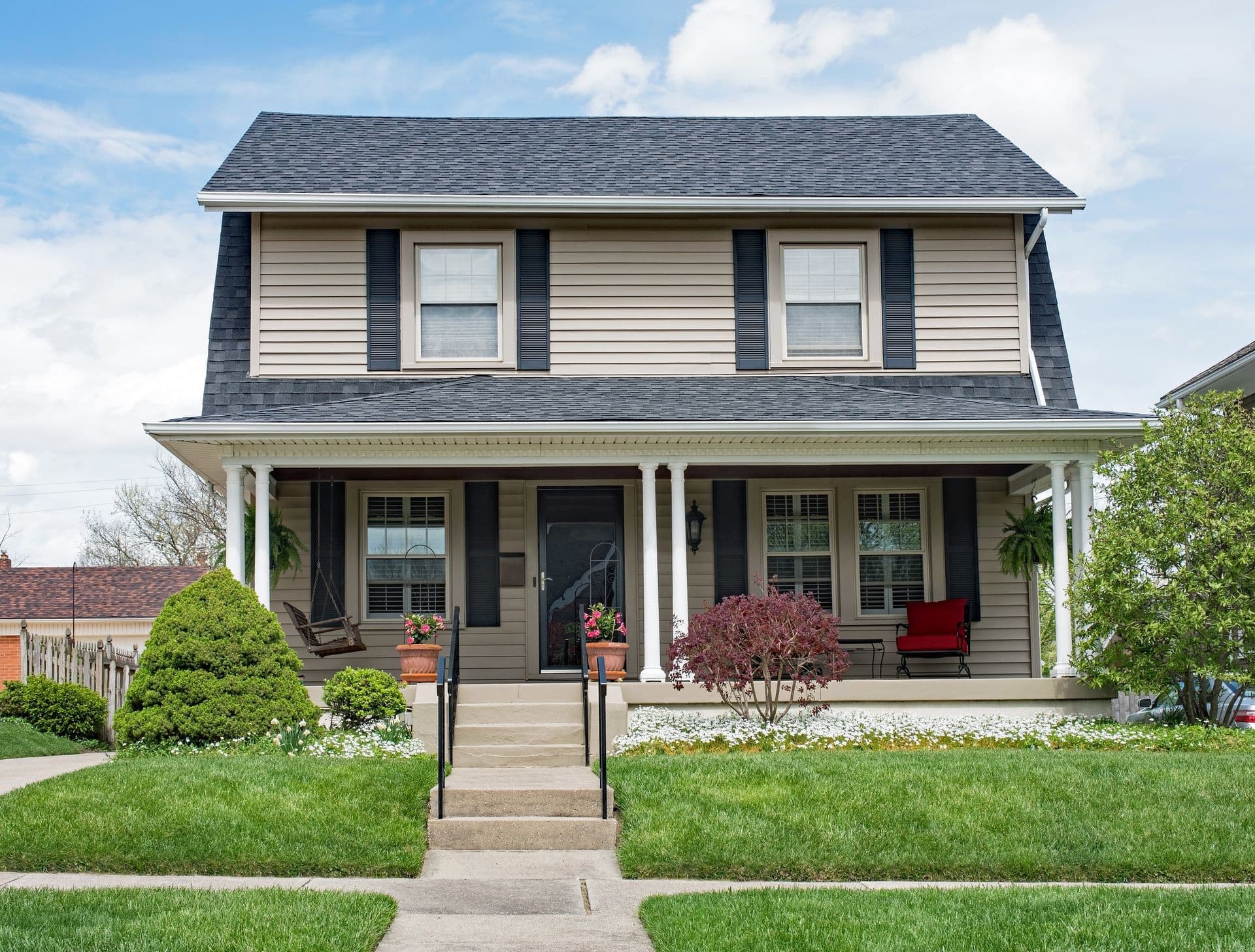 Two-story farmhouse with gray siding, dark shutters, and white front porch with columns, featuring manicured landscaping and flowering planters