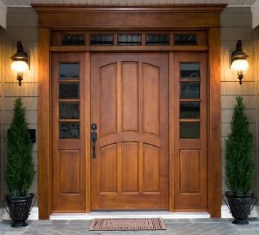 Wooden front entrance door with arched panels flanked by sidelights, wall sconces, and decorative potted plants