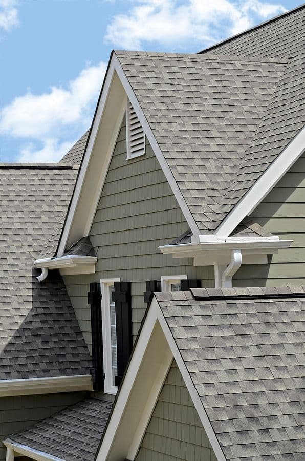 Residential house exterior showing gray asphalt shingle roofing, white trim, gable peaks with vents, and gutters against blue sky with clouds