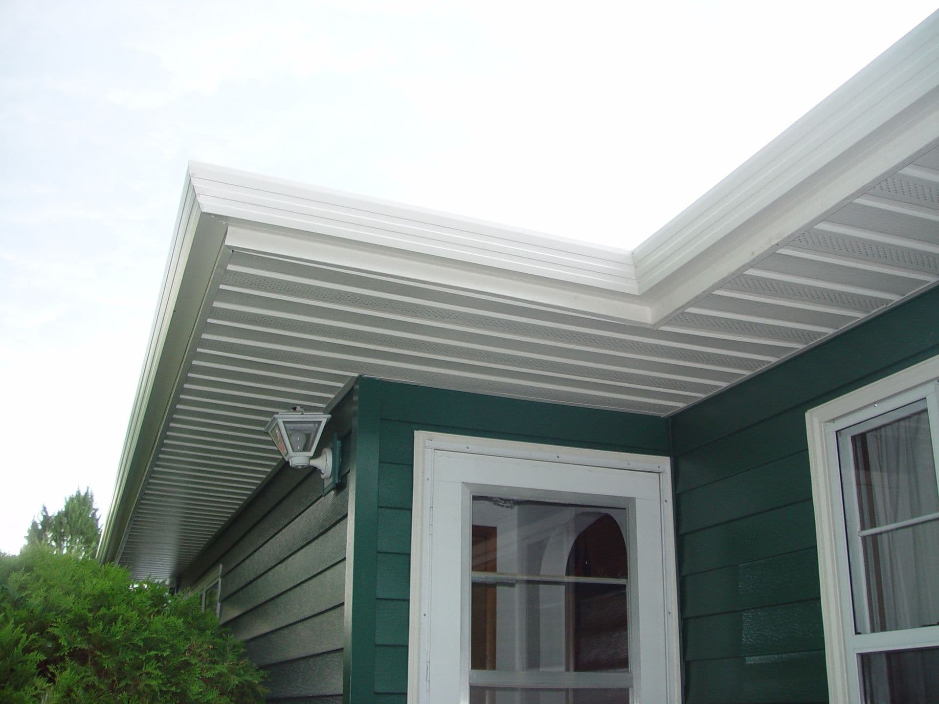 Retractable white aluminum awning over patio door and window of green-sided house, with wall lamp mounted on siding