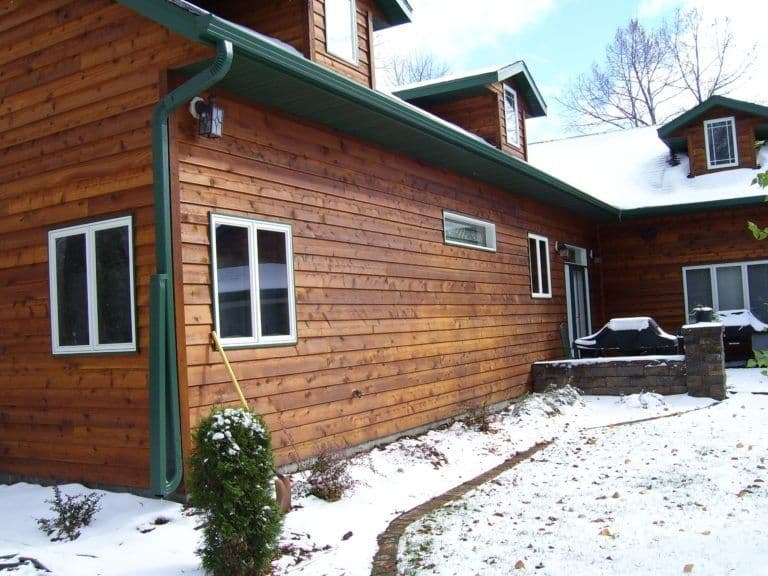 Exterior of a rustic log cabin with brown wood siding and green trim, featuring multiple windows and a gravel driveway with patches of snow, bare trees visible in background