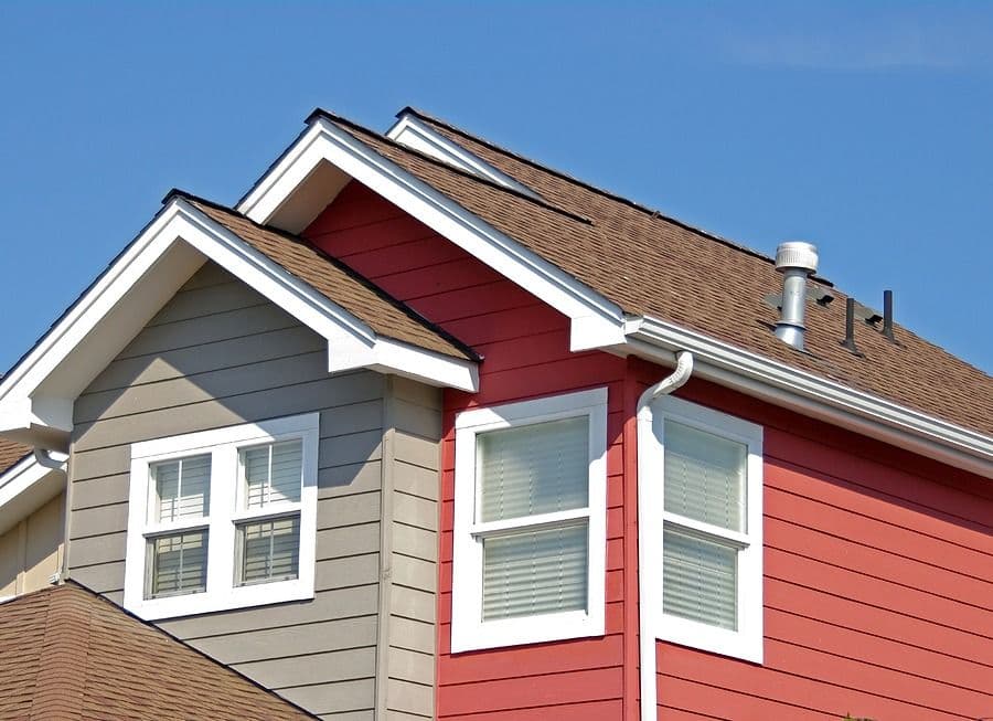 Modern house exterior showing two connected sections with gable roofs, one with tan siding and double windows, the other with red siding and tall double windows, brown shingle roof against clear blue sky