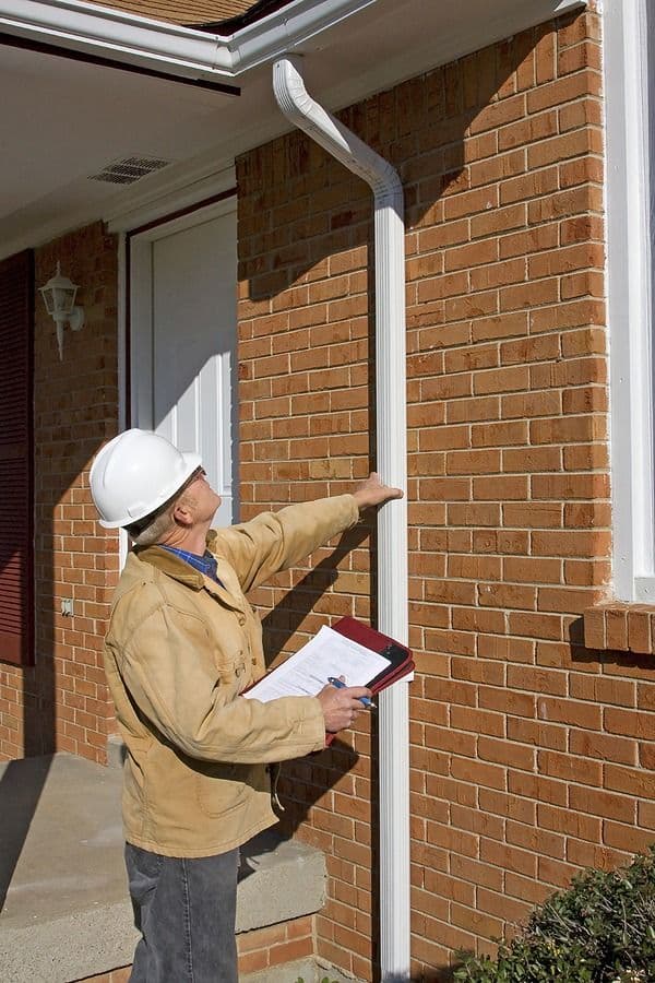 Home inspector in white hard hat and tan jacket examining a white downspout on brick exterior wall while holding clipboard