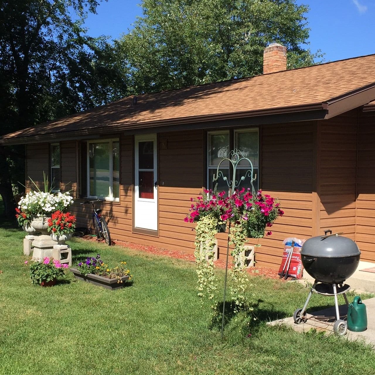 Brown single-story house with wood siding and brick chimney surrounded by landscaped yard featuring flowering plants, hanging baskets, grill, and bicycle under mature trees