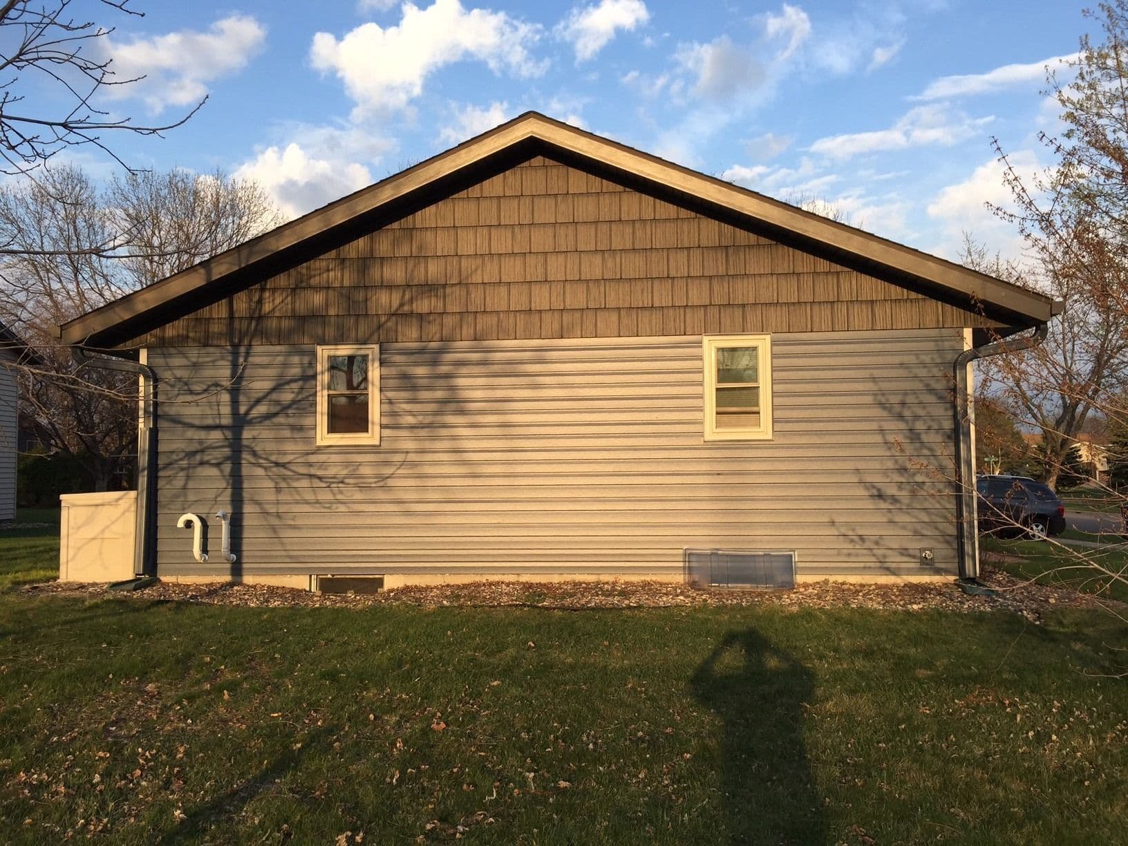 Single-story residential home with tan siding, two windows, gabled roof with wood shingles, and gravel foundation set against blue sky with bare trees
