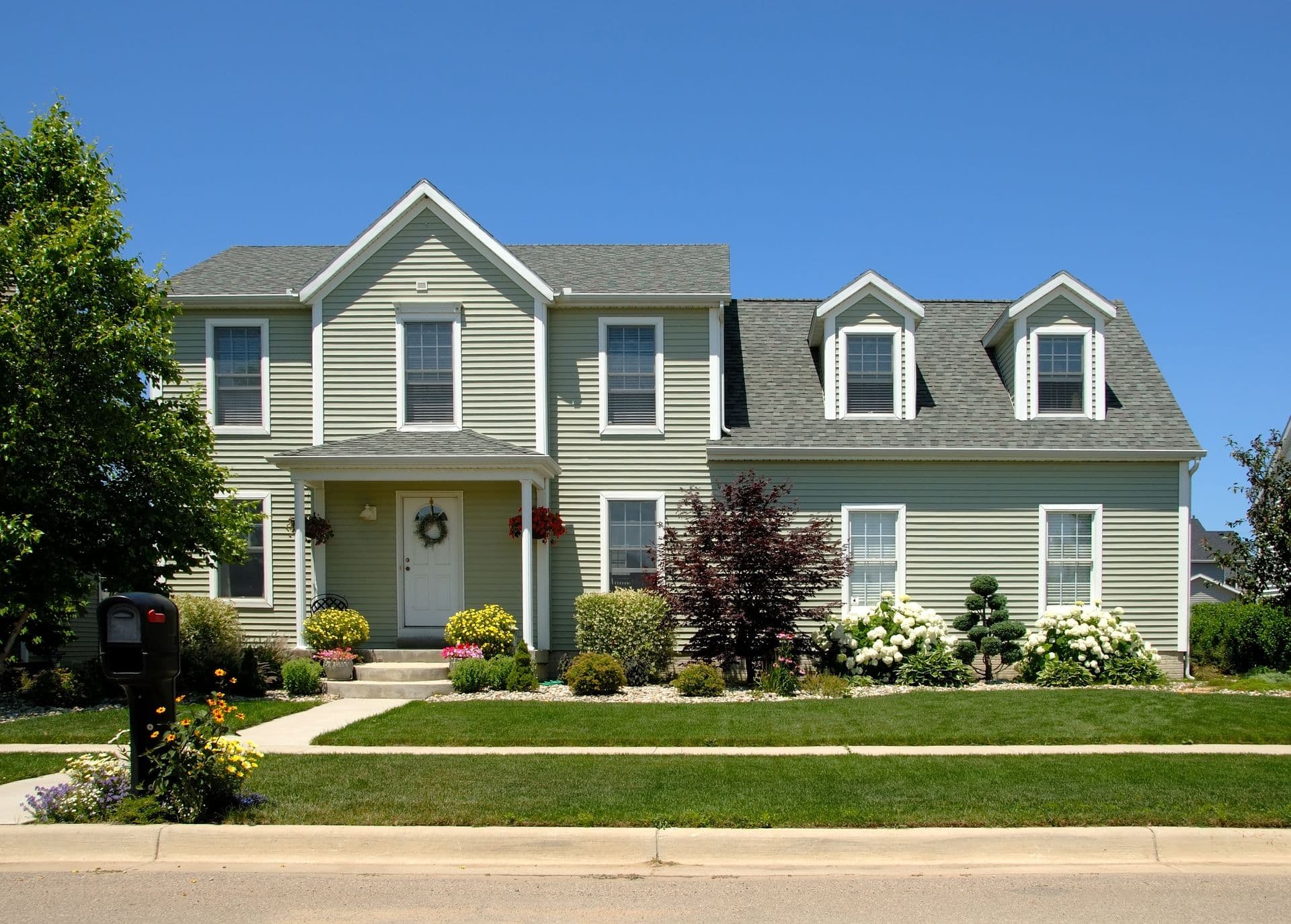 Two-story colonial-style home with pale green vinyl siding, white trim, and gray roof, featuring manicured landscaping with flowering shrubs and green lawn