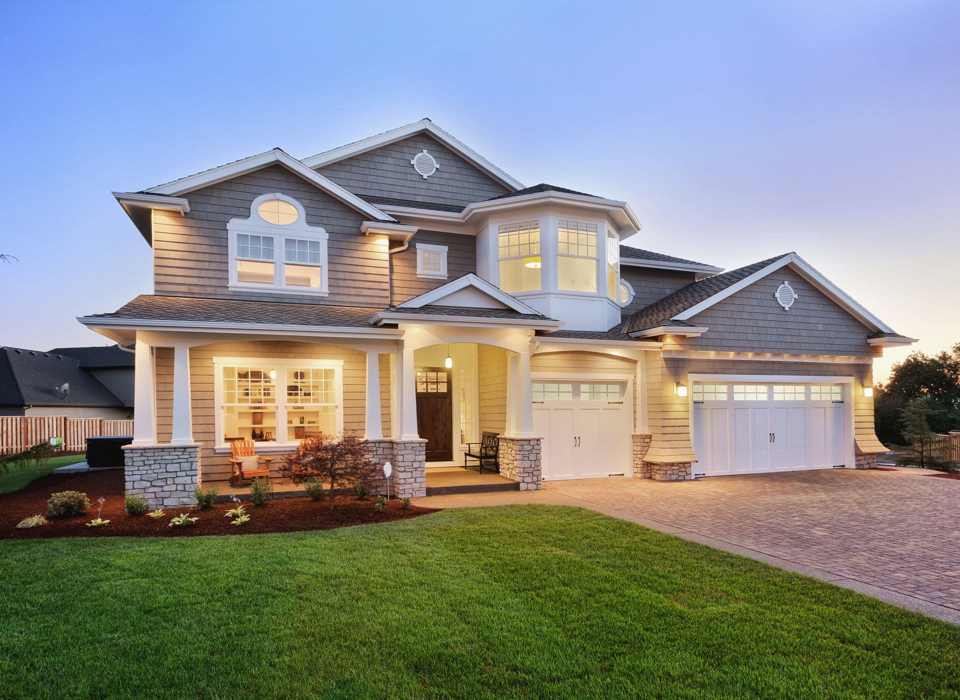 Modern two-story home at dusk with white columns, stone accents, dual garage doors, illuminated windows, manicured lawn, and brick driveway
