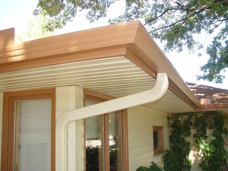 Modern home exterior showing wooden pergola with cream-colored metal siding and curved gutter, featuring glass doors and climbing ivy on light-colored wall