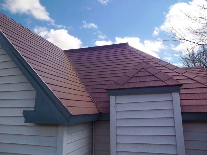 Burgundy metal roof with multiple peaks and angles on residential house with white siding and dormers, against blue sky with clouds