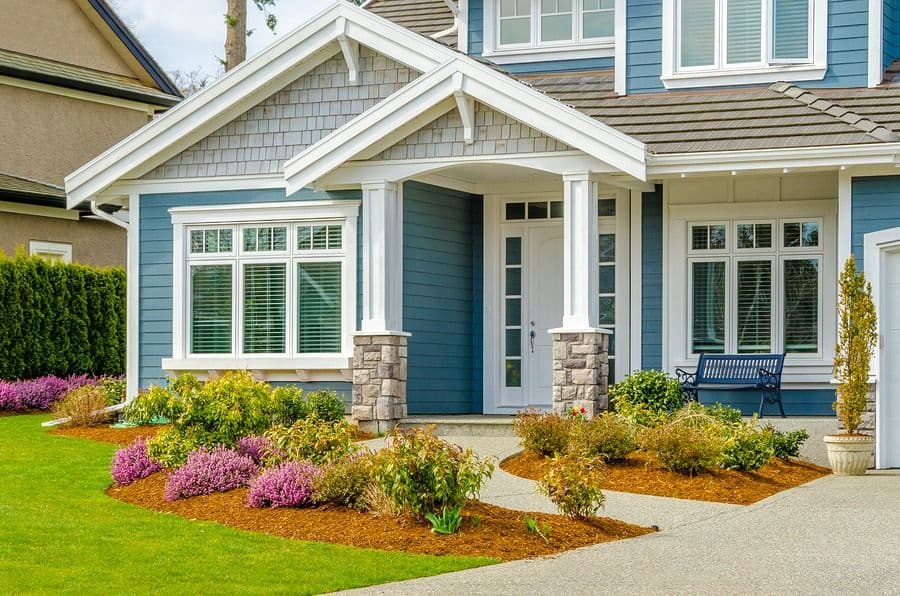 Blue contemporary home with covered front porch, white trim, and stone columns, featuring landscaped garden with pink flowering shrubs and mulch beds along the driveway