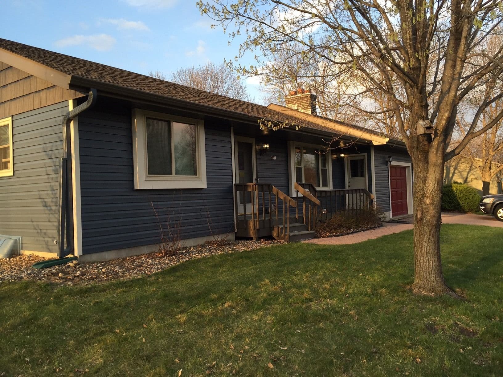 Single-story blue ranch home with front porch, brick chimney, and attached garage, surrounded by bare trees and lawn