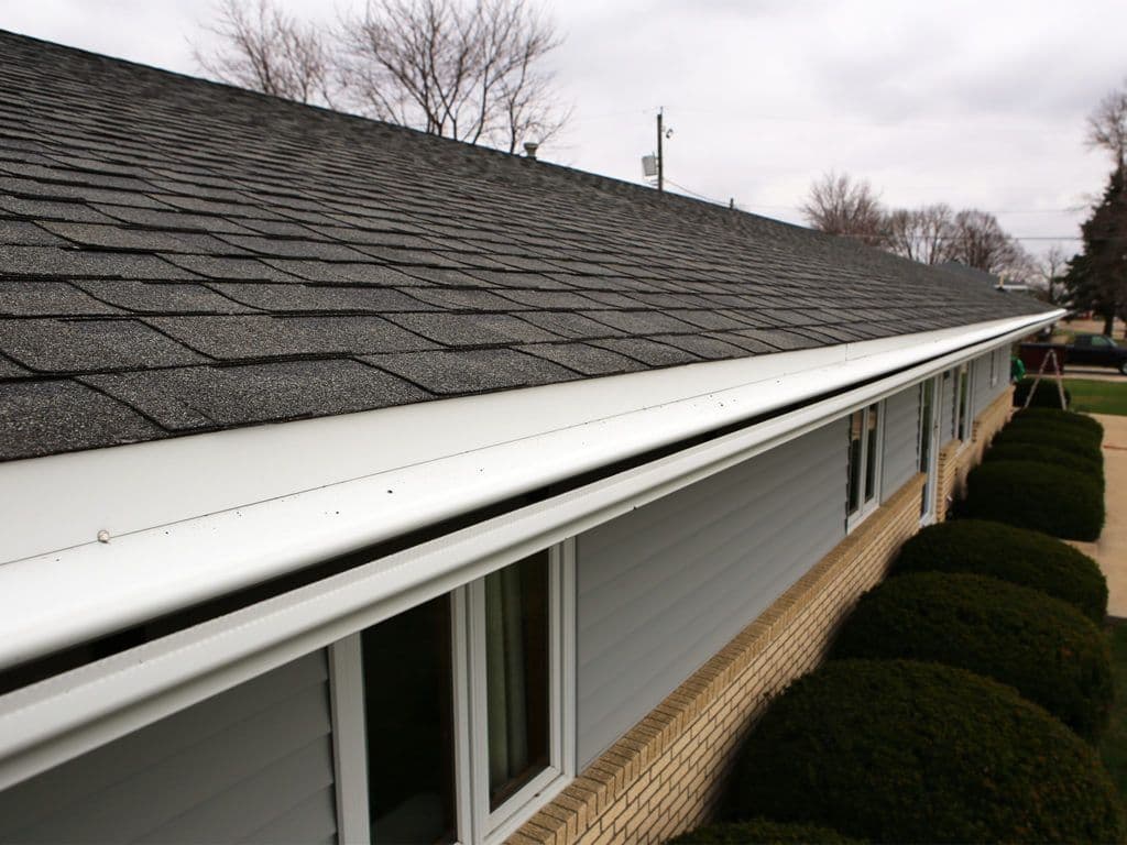 Residential roof with dark asphalt shingles and white gutters running along the roofline of a brick house