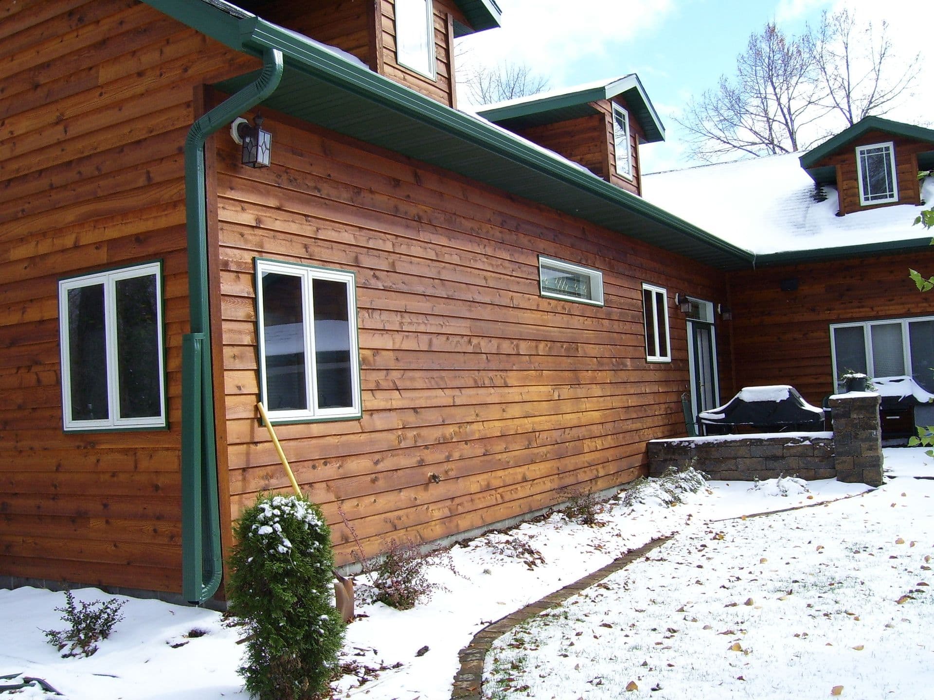 Log cabin exterior with green metal roof and gutters, multiple white-trimmed windows, stone foundation, gravel driveway, and evergreen landscaping with patches of snow
