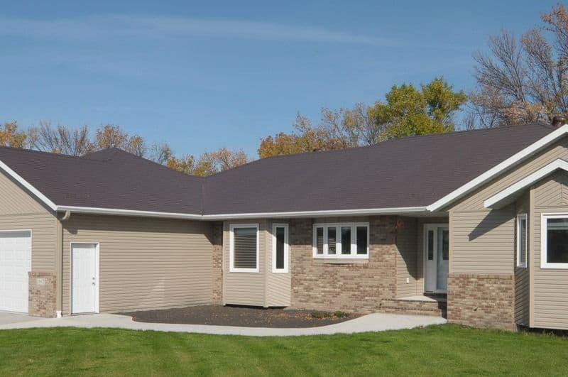 Modern single-story ranch home with tan siding, brick accents, dark roof, attached garage, and manicured lawn with autumn trees in background