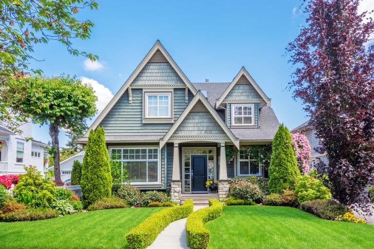 Modern two-story home with gray siding and gabled roofline, manicured lawn with curved walkway, evergreen shrubs, and flowering plants under clear blue sky