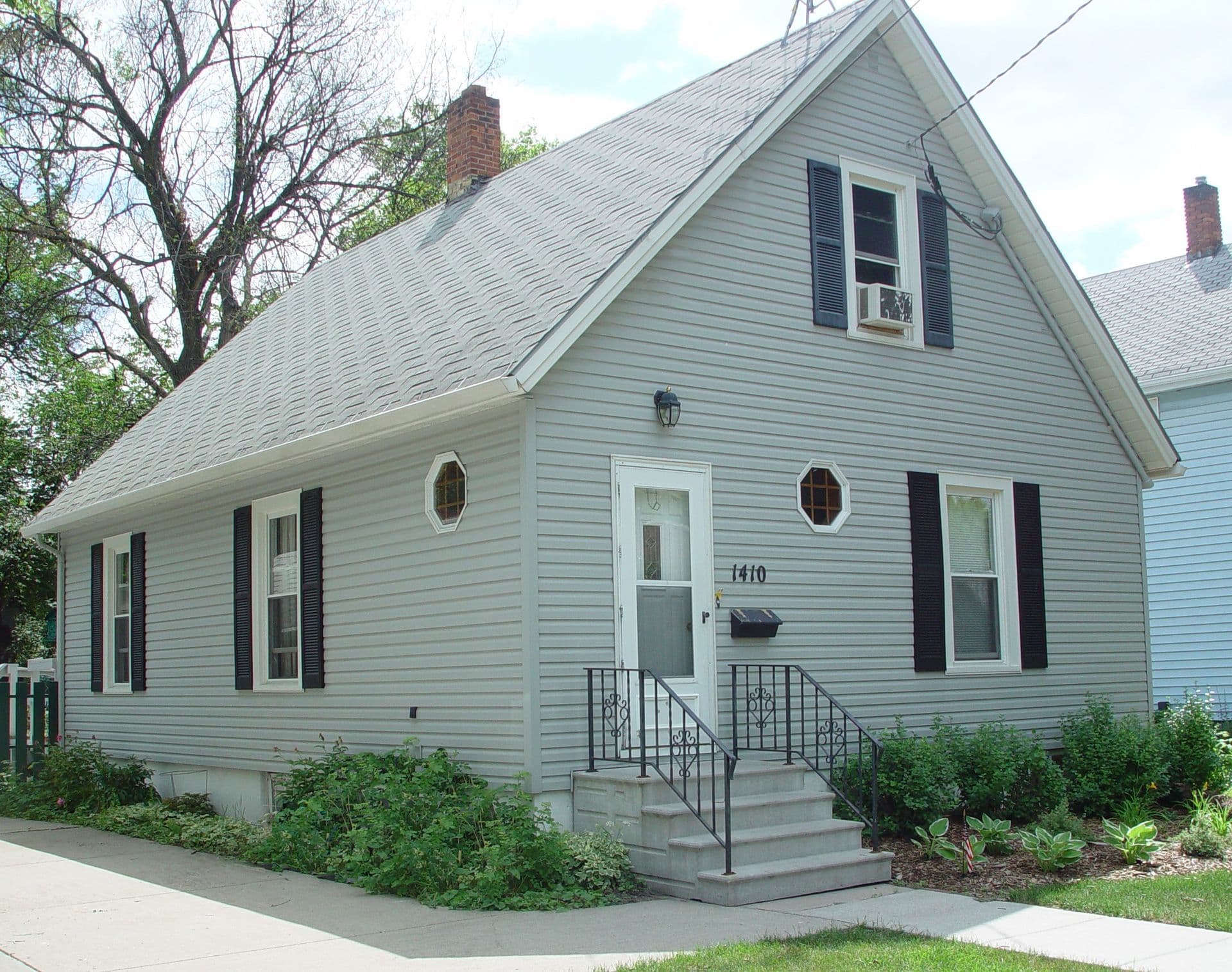 Light blue single-story house with gabled roof, brick chimneys, white front door marked 1410, black shutters, and metal porch railing surrounded by landscaping