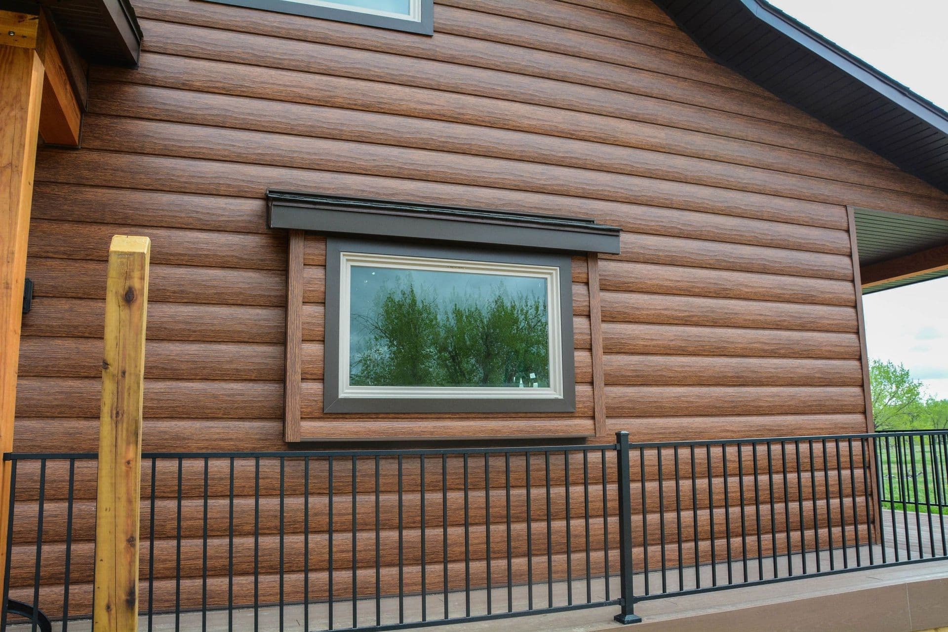 Log cabin exterior with horizontal wood siding, metal-framed window reflecting evergreen trees, and black metal deck railing