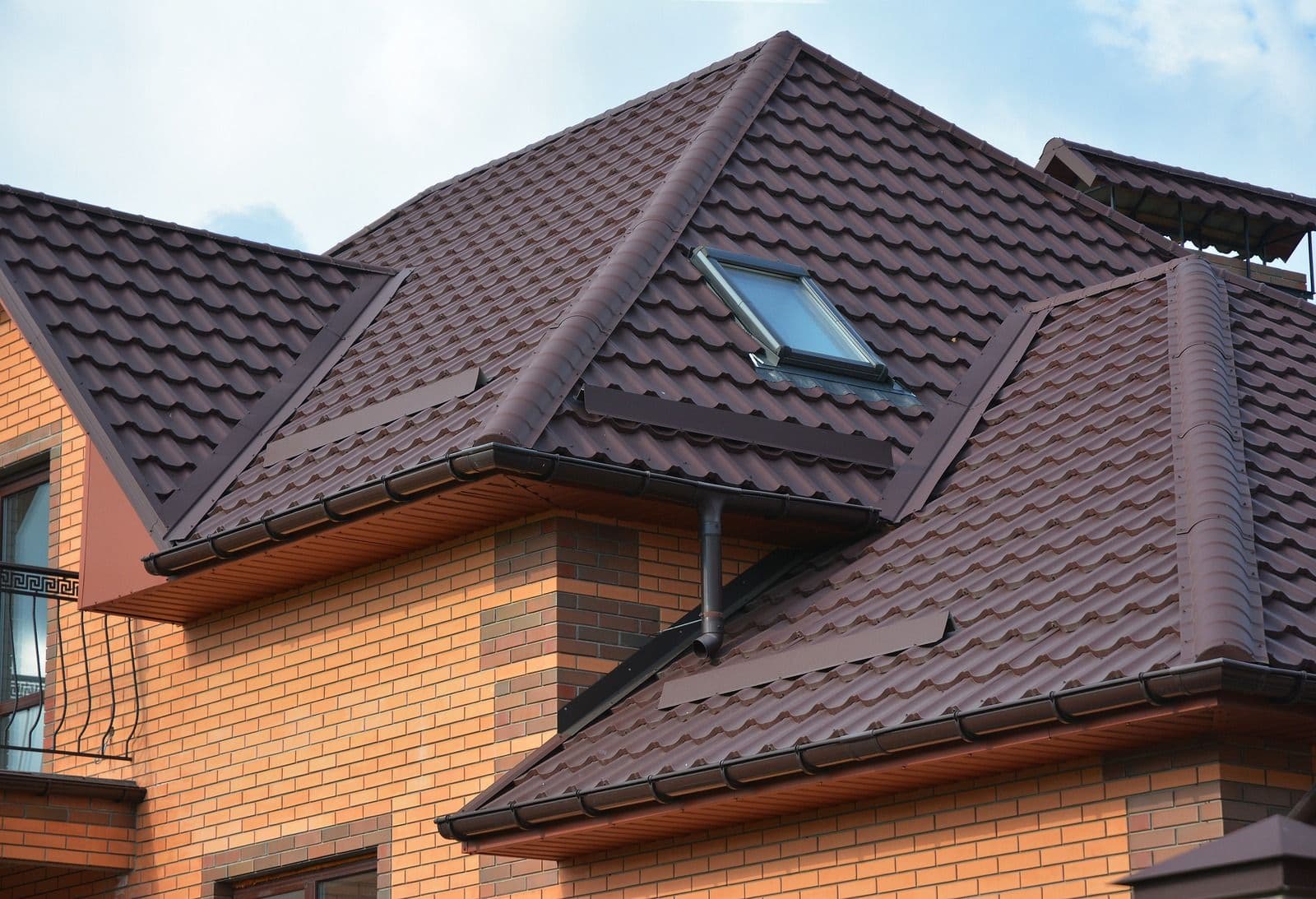 Modern residential roof with burgundy metal shingles, dormer window, and dark guttering on red brick house exterior