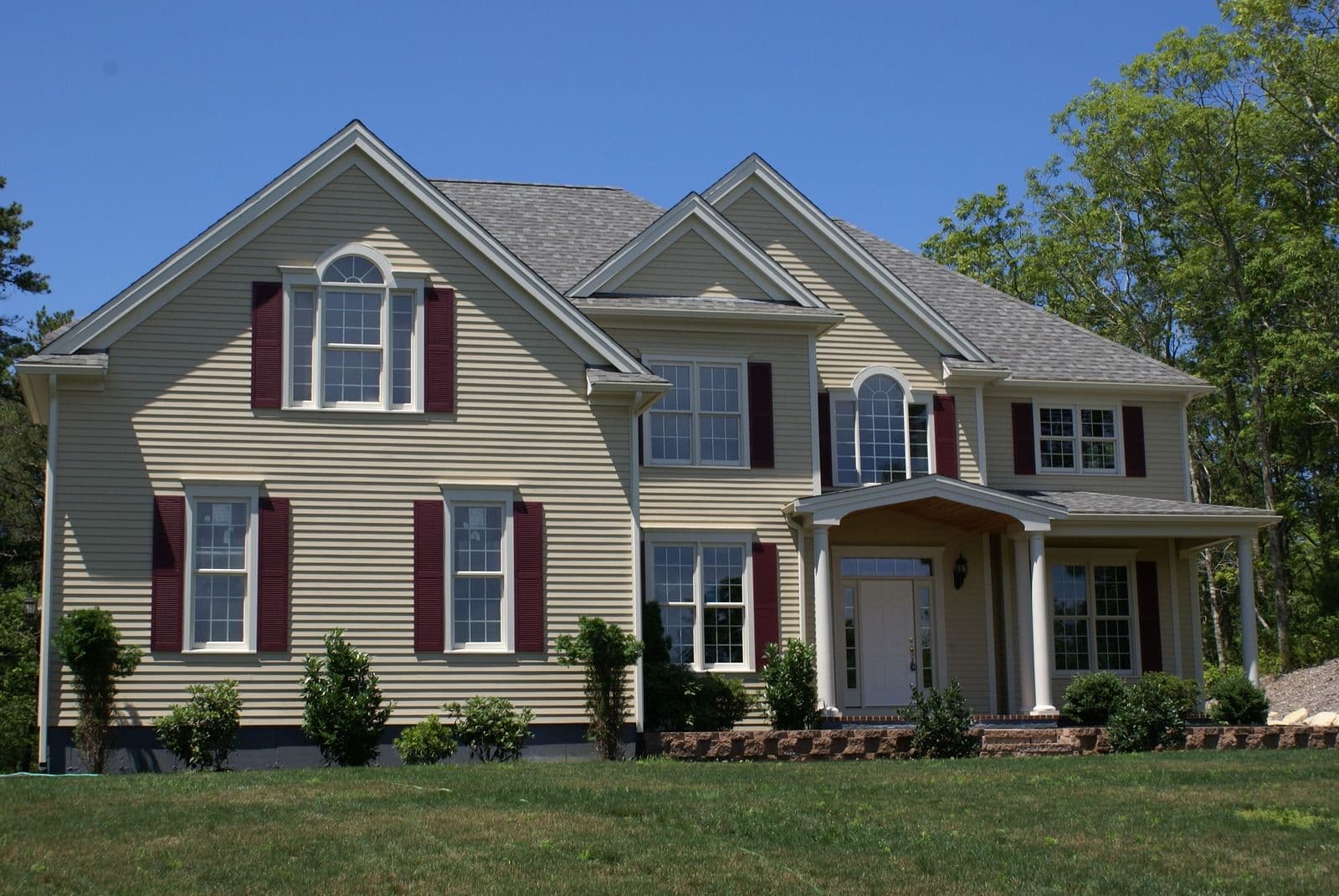 Two-story suburban home with tan siding, red shutters, gabled rooflines, front portico with columns, multiple windows, and landscaped lawn