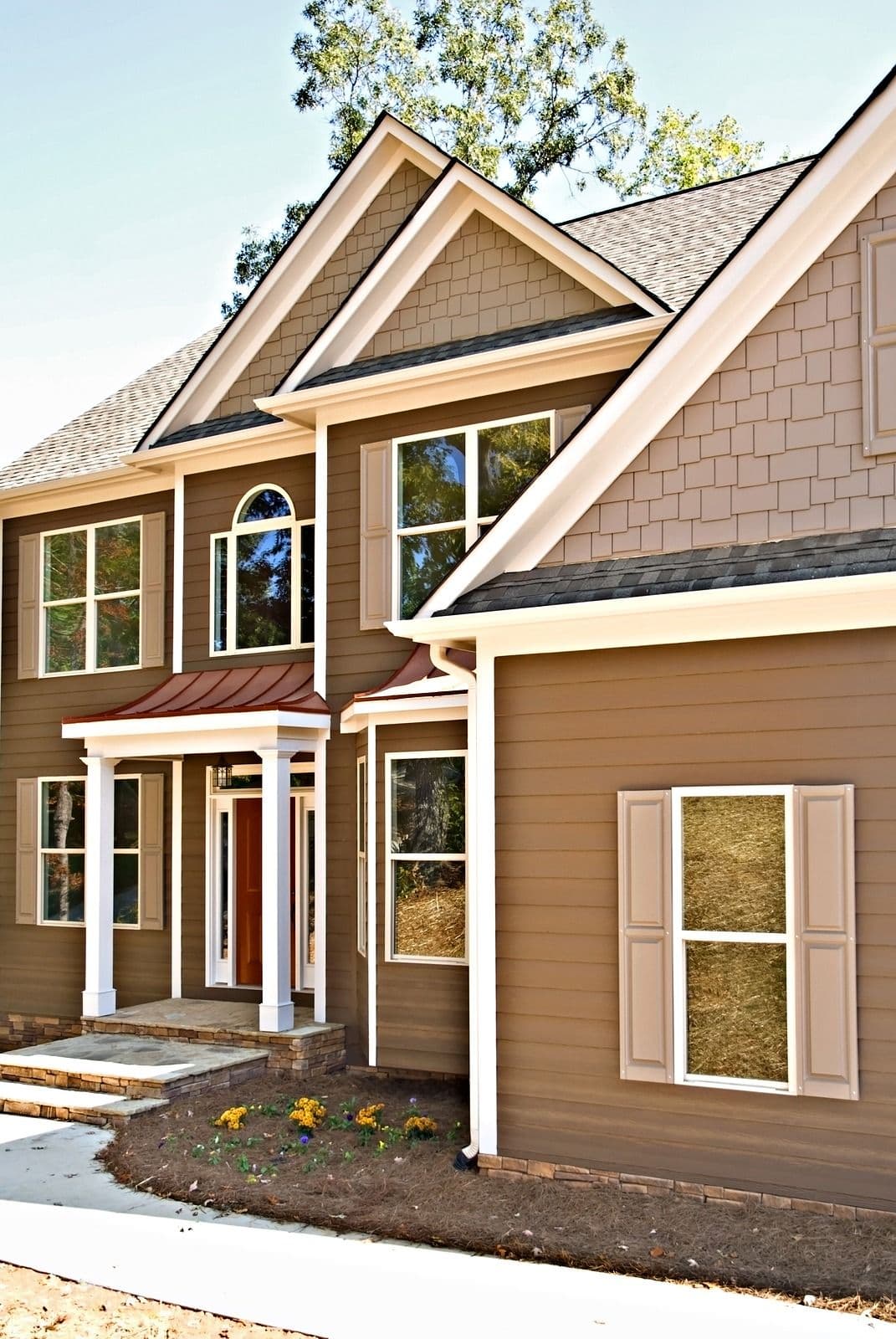 Front exterior of a two-story brown and tan house with white trim, front porch, gabled roof, multiple windows, and garage
