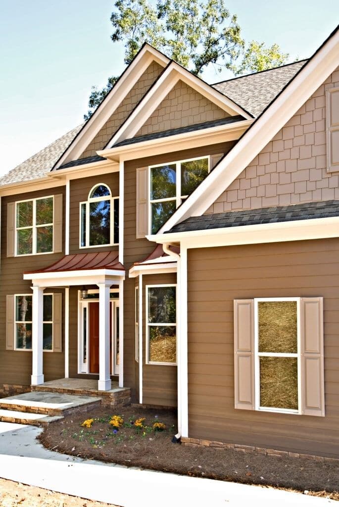 Two-story brown house with white trim, covered front porch, and attached garage, featuring multiple windows and landscaping with yellow flowers
