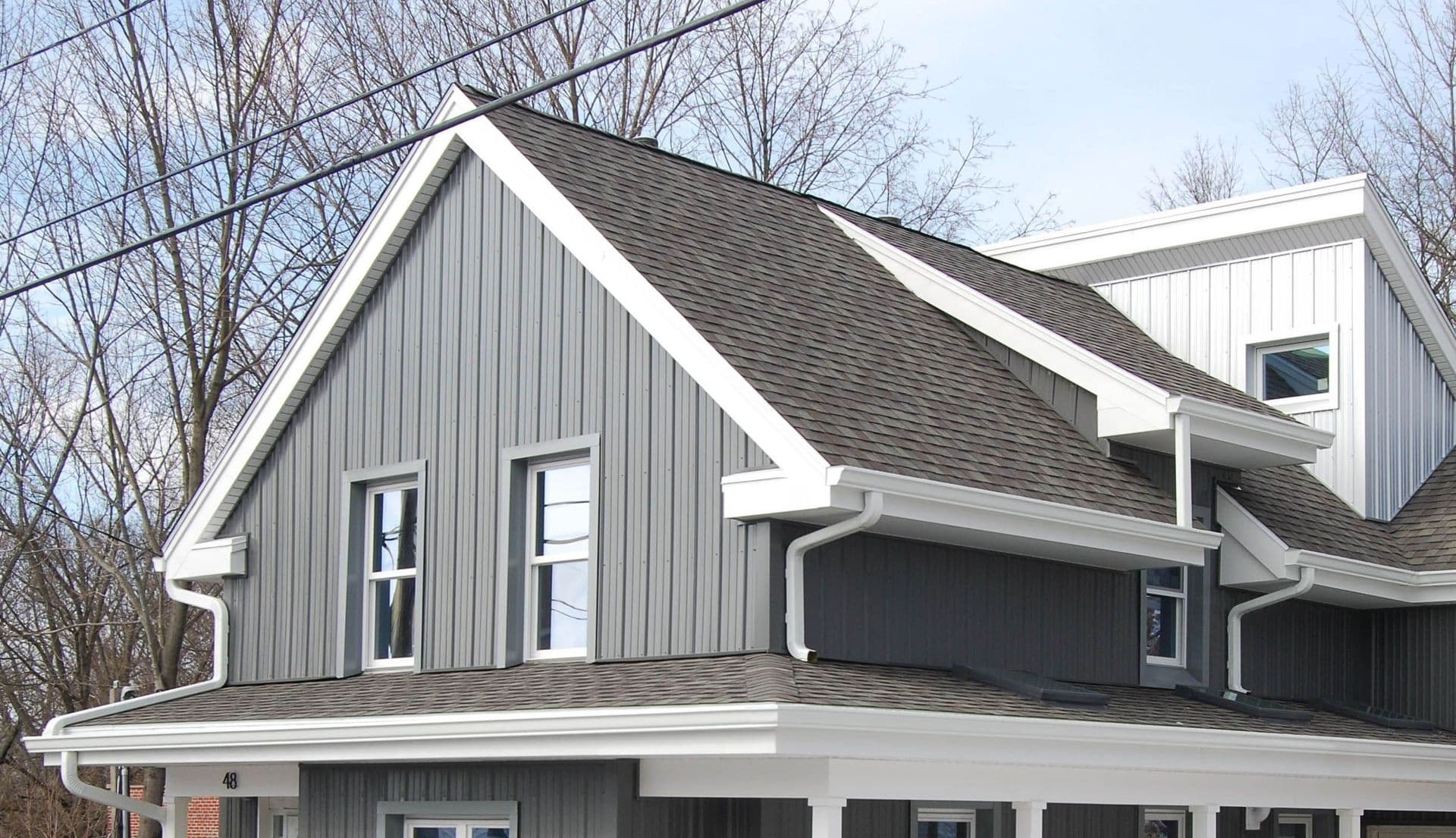 Modern two-story house with gray metal siding, white trim, dark shingle roof, and attached garage in winter setting surrounded by bare trees