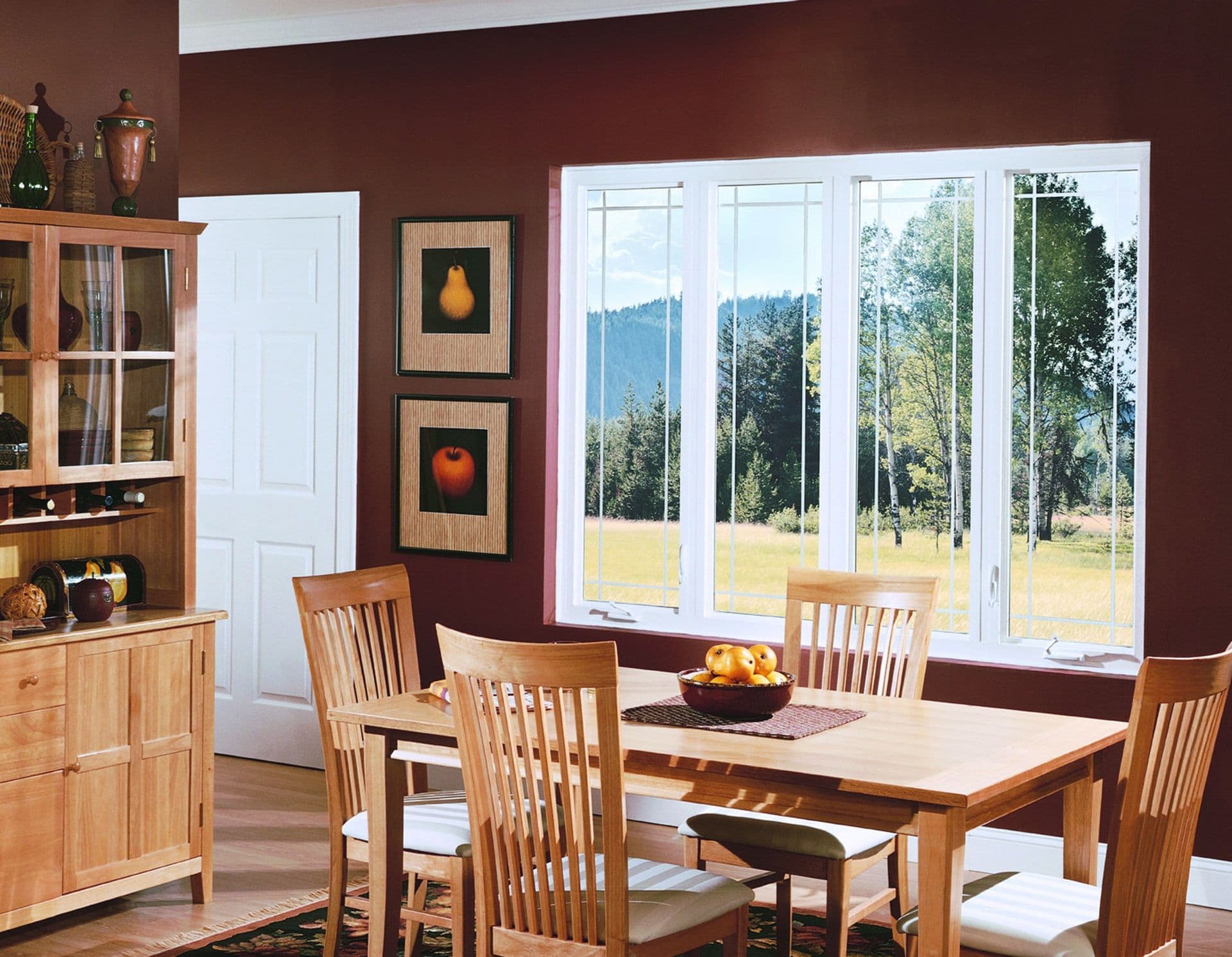 Dining room with wood furniture, burgundy walls, large multi-pane windows overlooking mountains and fields, fruit artwork, and built-in cabinetry