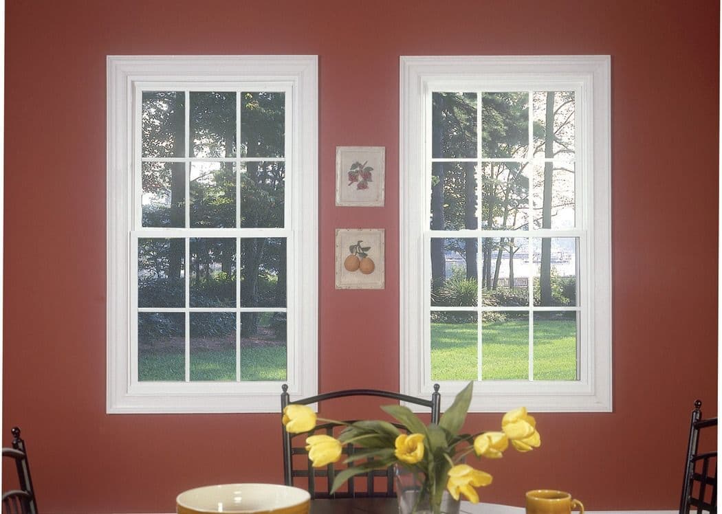 Two white-framed windows on a terrace wall with forest views, botanical prints between them, and yellow tulips in a vase on the table below