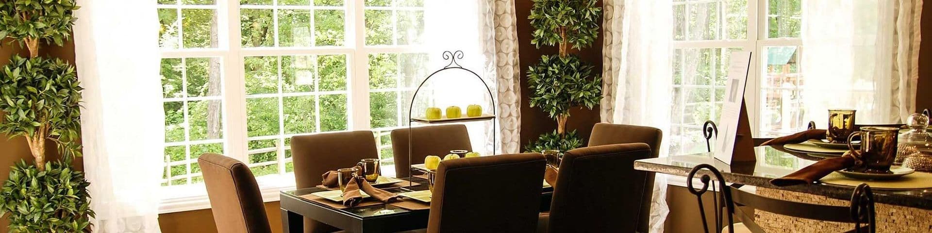 Elegant dining room with brown upholstered chairs around a black table, large windows overlooking greenery, potted plants, and decorative accessories in warm gold and bronze tones