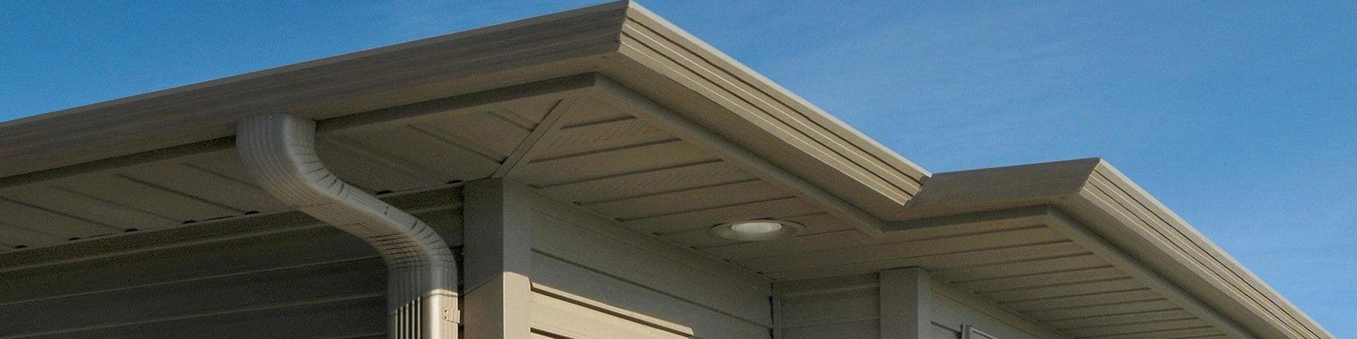 Modern residential building exterior with metal roof overhang, downspout, and recessed lighting under clear blue sky