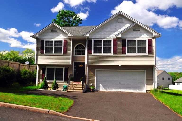 Two-story suburban house with cream siding, red shutters, two-car garage, front porch, and manicured lawn under blue sky with clouds