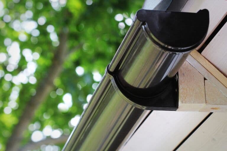 Metallic gutter and downspout attached to white fascia board and wooden soffit, with green foliage in background