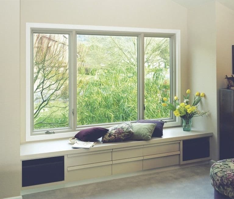 Built-in window seat with cushions and decorative pillows beneath triple-pane windows overlooking green garden, with yellow flowers in vase on sill